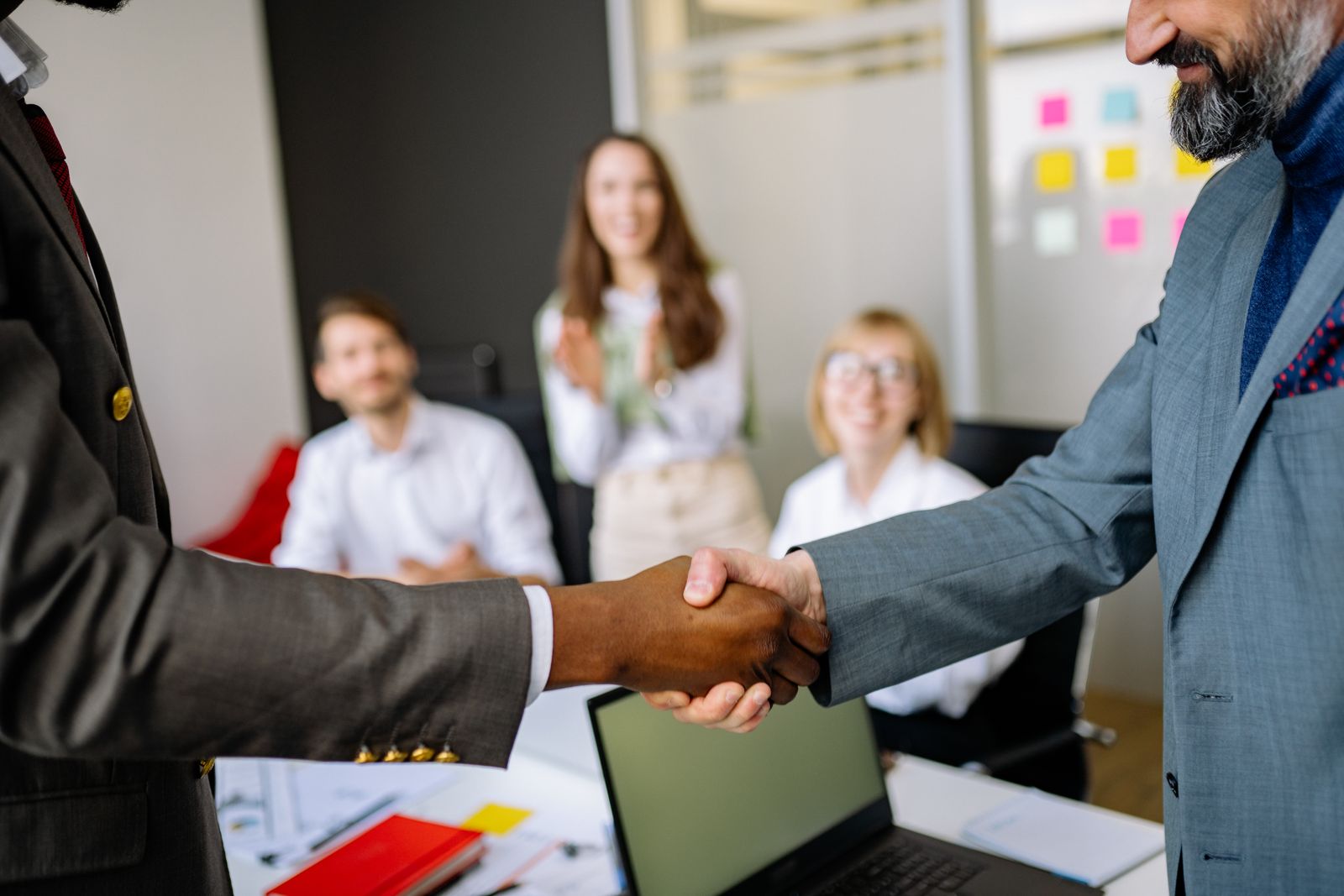 Two colleagues shaking hands in an office while coworkers look on